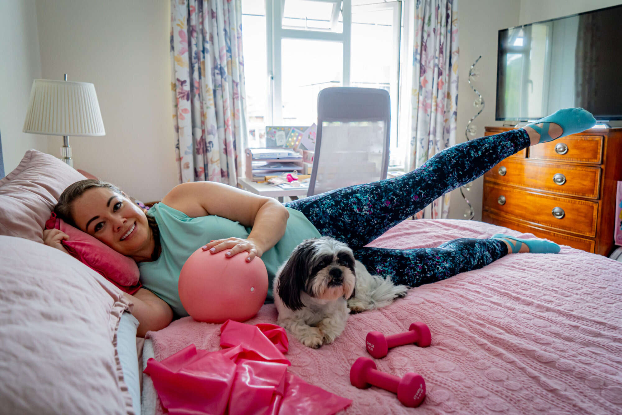 Leanne lying on her side on her bed, leg raised, resting a hand on a pink ball, with Stanley her dog nearby.