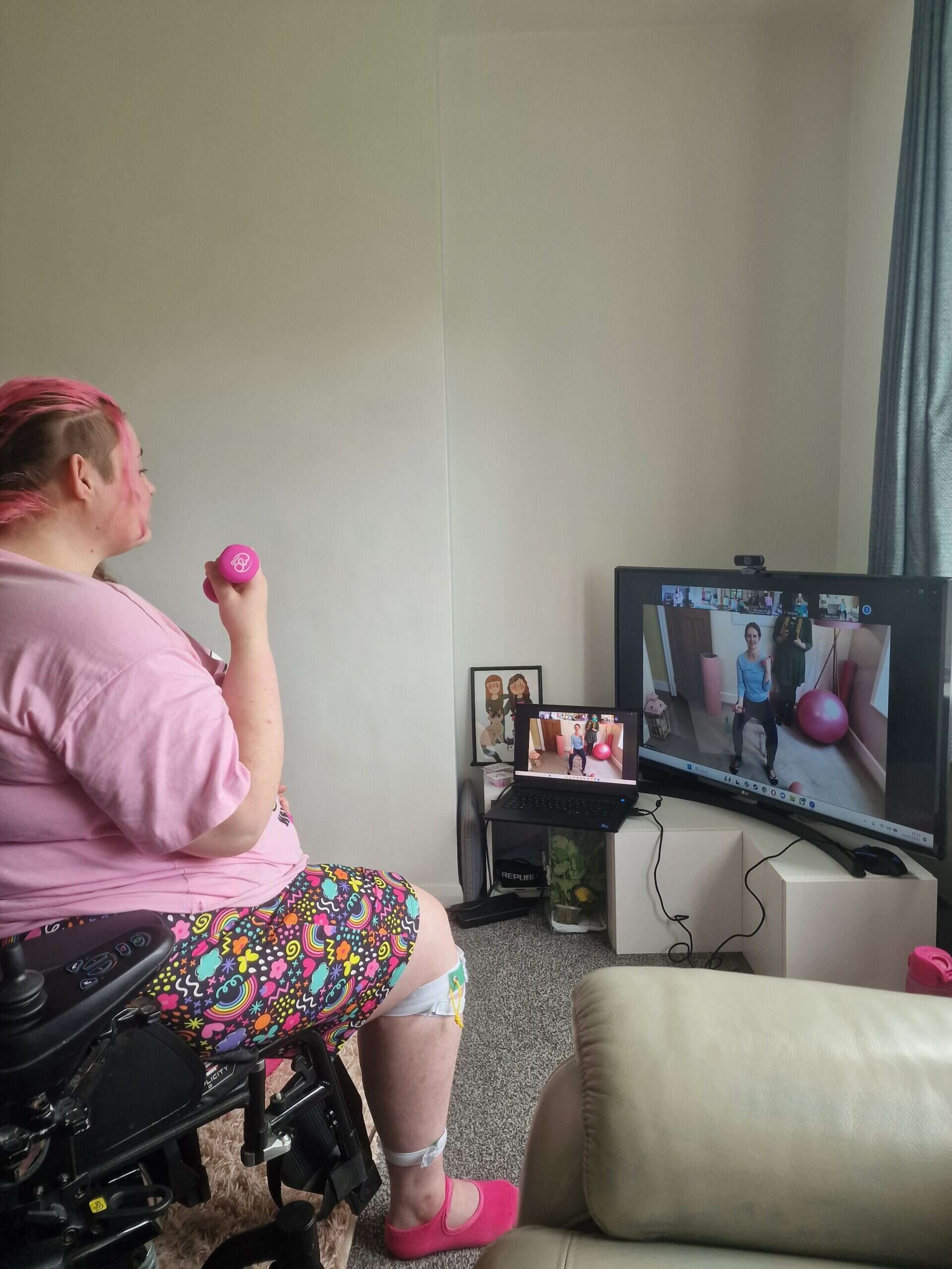 Our member Sam in her wheelchair holding pink weights, with teacher Kerry demonstrating a move on screen.