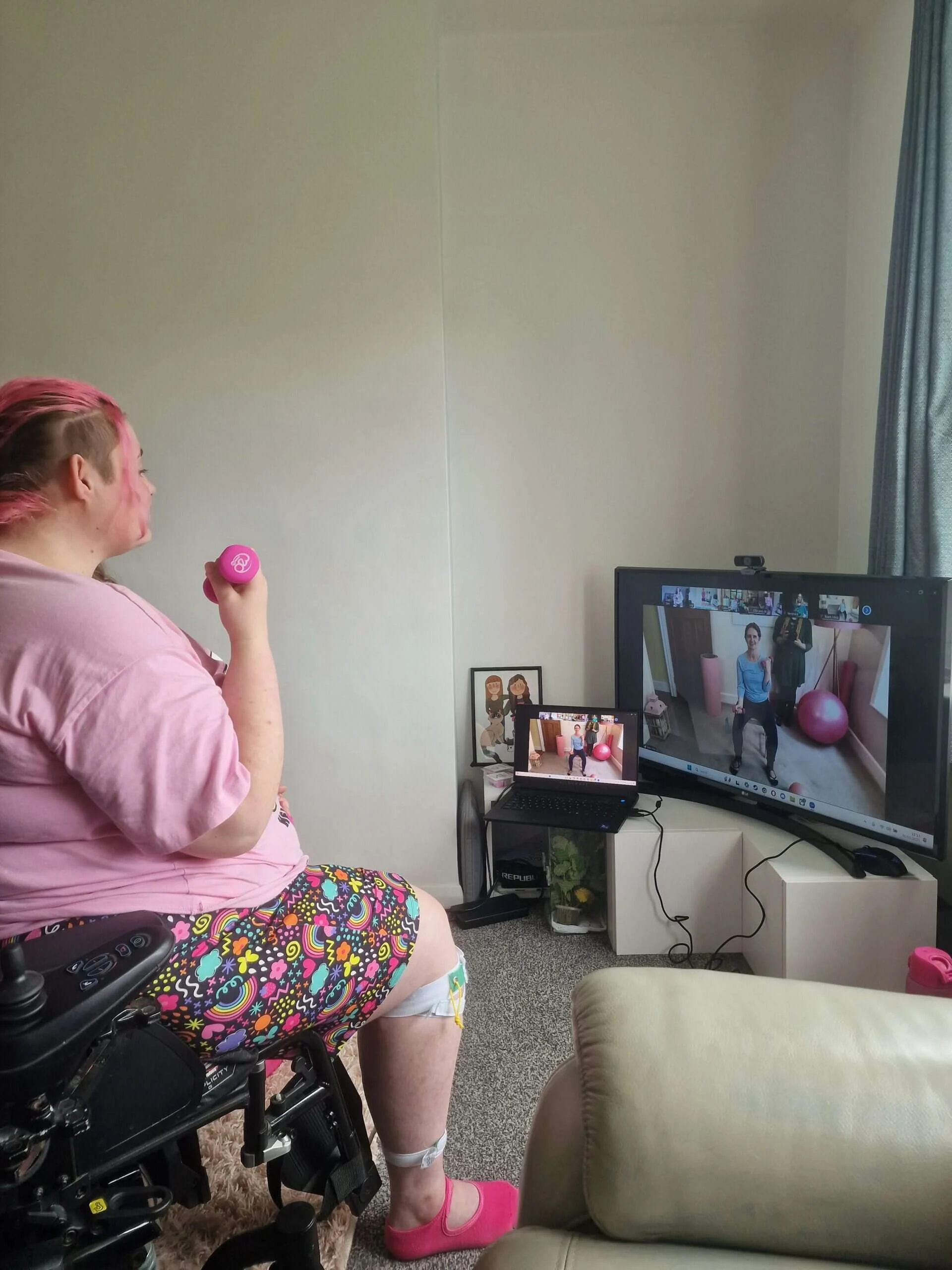 Our member Sam in her wheelchair holding pink weights, with teacher Kerry demonstrating a move on screen.