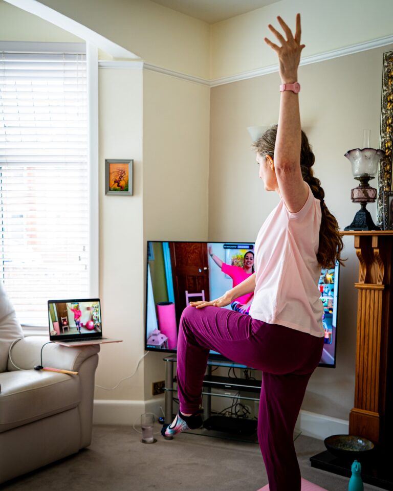Kerry, in pink workout wear, balancing on one leg while tapping her knee and reaching up.