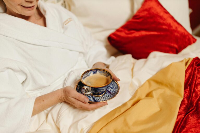 A woman relaxing in bed with a cup of tea, wrapped in red velvet and gold covers and cushions.
