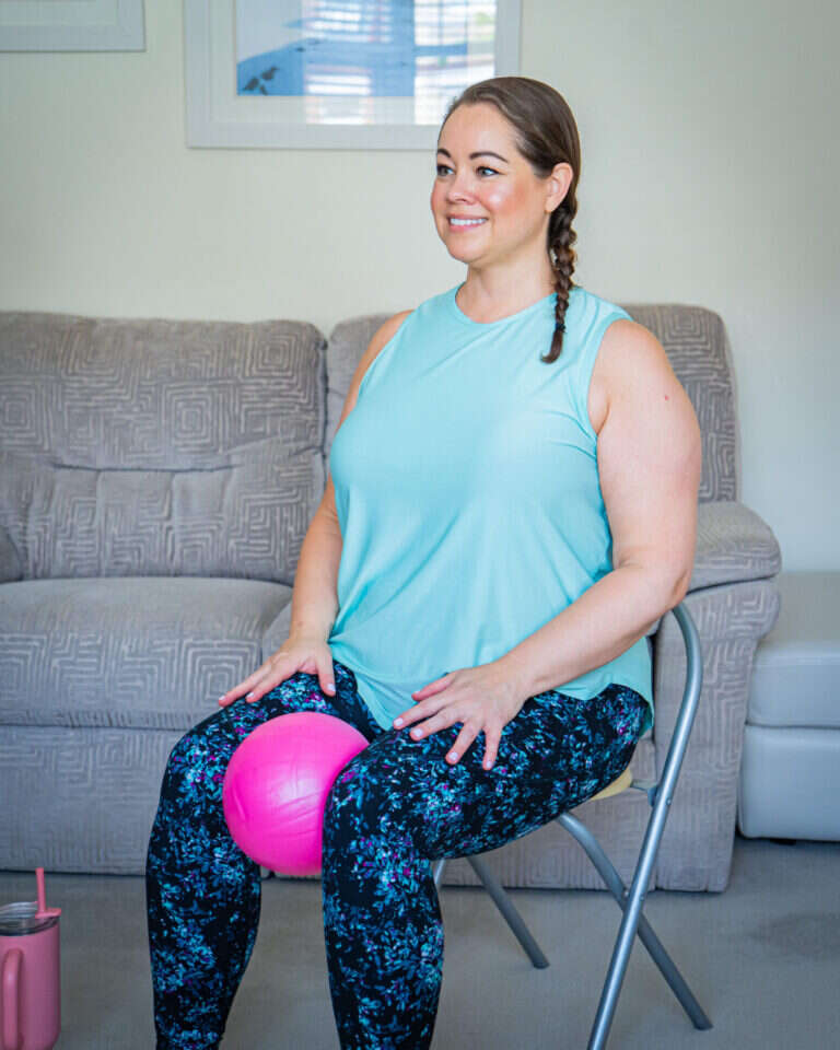 Leanne sitting on a chair wearing a sleeveless turquoise top and multicloured leggings. Holding a pilates ball between her knees.