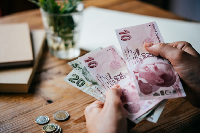 Two hands holding and counting British pound coins and notes, with a desk, vase and notebooks in the background.