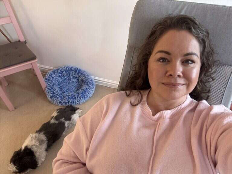 Leanne sitting comfortably in a pink sweater, smiling, with a small black-and-white dog resting beside her.
