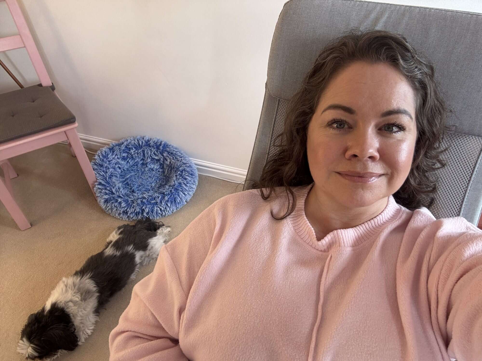 Leanne sitting comfortably in a pink sweater, smiling, with a small black-and-white dog resting beside her.
