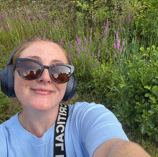 Chloe taking a selfie outdoors with greenery and purple heather behind her, smiling.