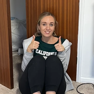 Martha sitting on the floor in a black outfit and grey hoody, giving a thumbs up and smiling.