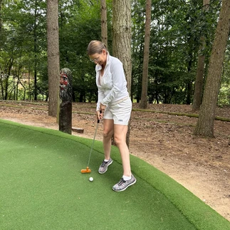 Suzanne playing mini golf, standing and smiling, about to hit the ball with her golf club.