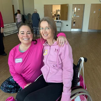 Leanne crouching beside Suzanne's wheelchair, which has pink flowers on the wheels. Both are smiling.