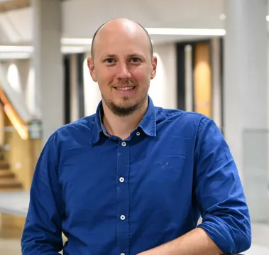 Alex, a white male wearing a blue shirt with white buttons, smiling at the camera.