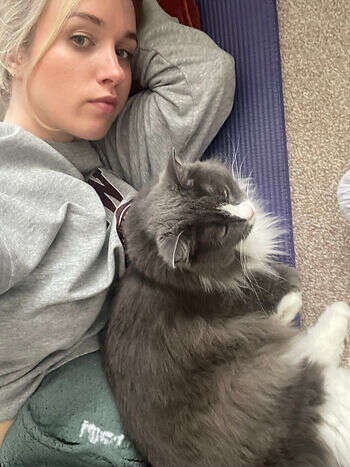 Martha taking a selfie with her fluffy grey-and-white cat, both lying on her purple Pilates mat.