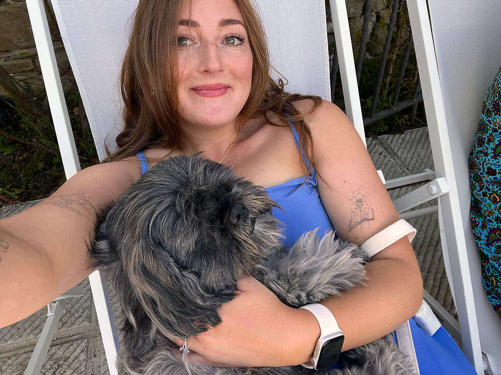 Chloe sat on a white deck chair with a fluffy grey dog on her lap, wearing a blue dress and smiling.