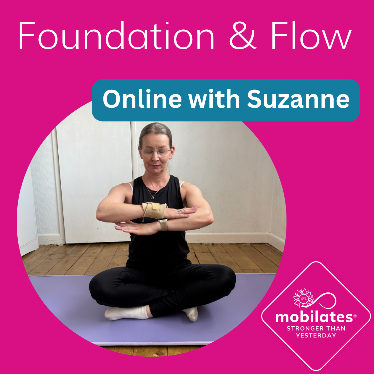 Suzanne seated cross-legged on a yoga mat, wrists crossed in front of her chest in a seated stretch, in a home studio setting