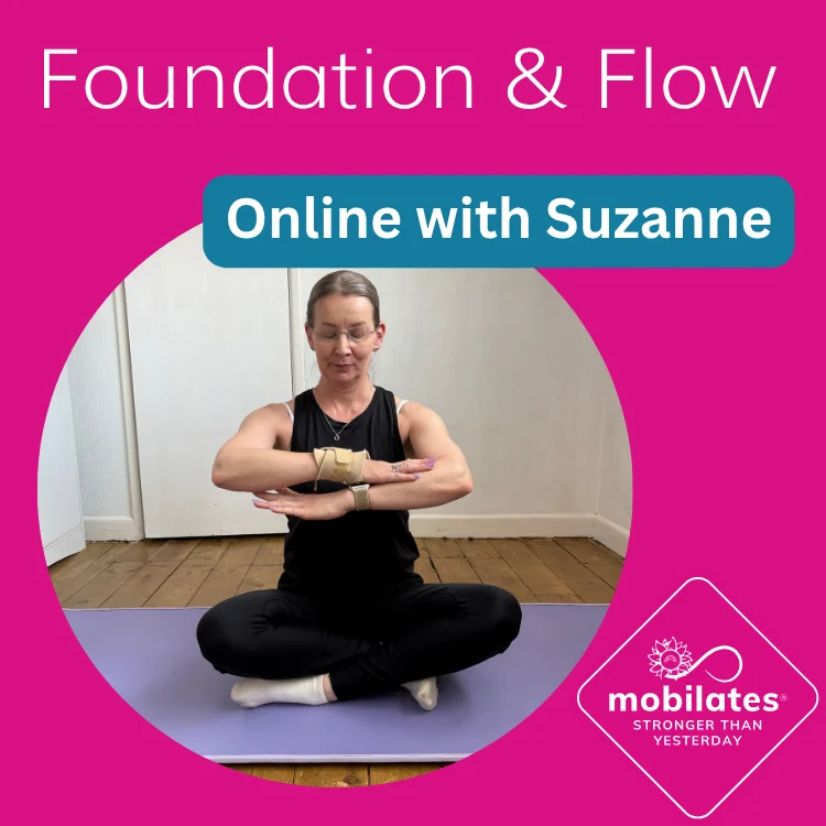 Suzanne seated cross-legged on a yoga mat, wrists crossed in front of her chest in a seated stretch, in a home studio setting