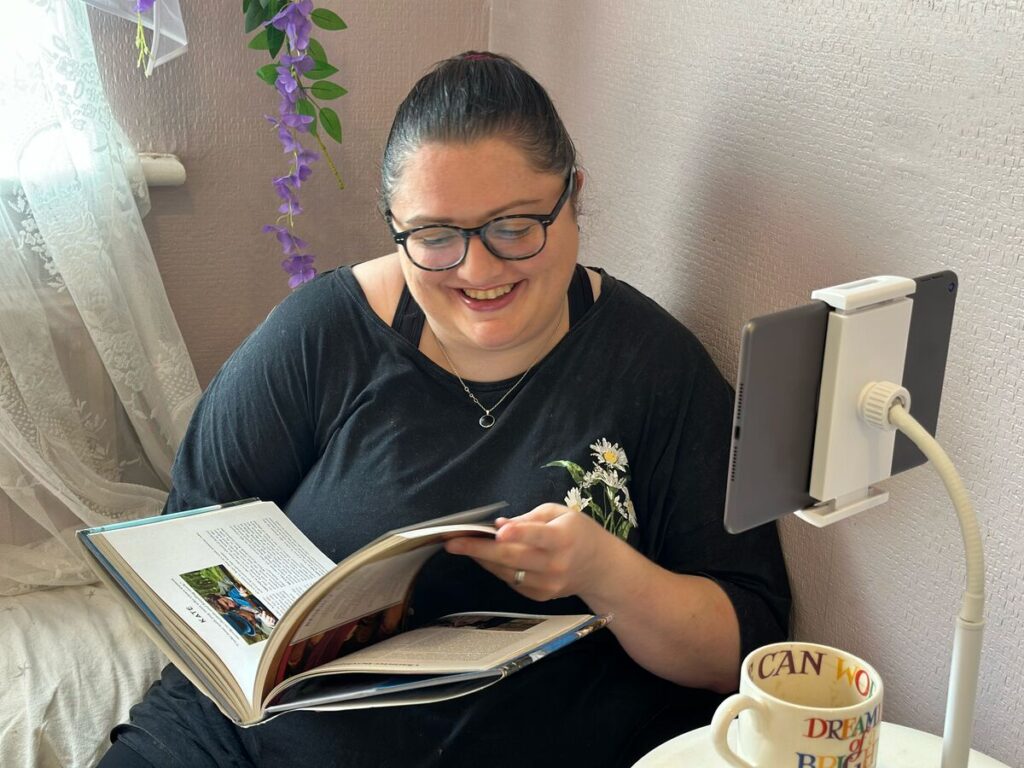 A woman smiles as she reads a book at home, with a mug beside her and a tablet set up for an online session.
