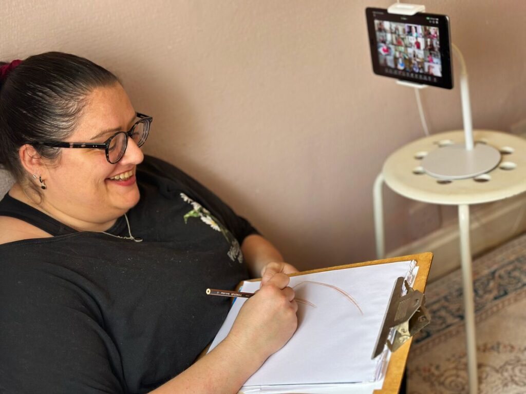 A woman draws on a clipboard at home while taking part in an online session on a tablet.