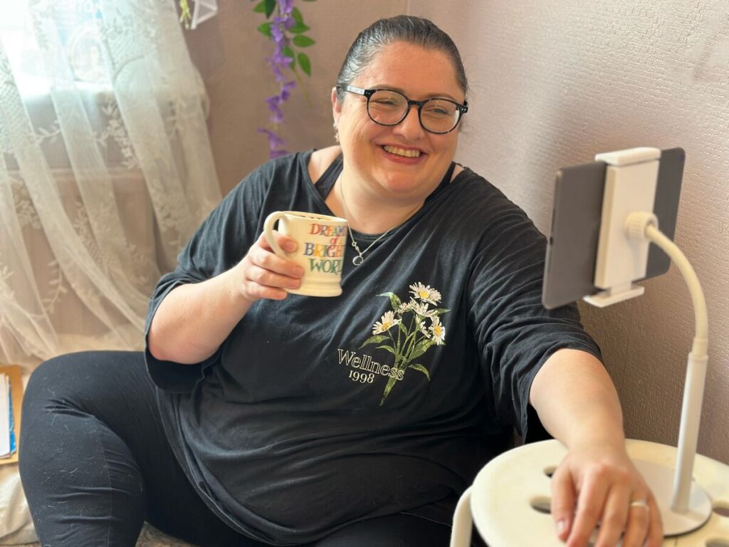 A smiling woman sits at home with a mug, chatting to an online session on a tablet.