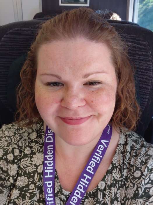 Rachel smiling on a train, wearing a purple Verified Hidden Disabilities lanyard