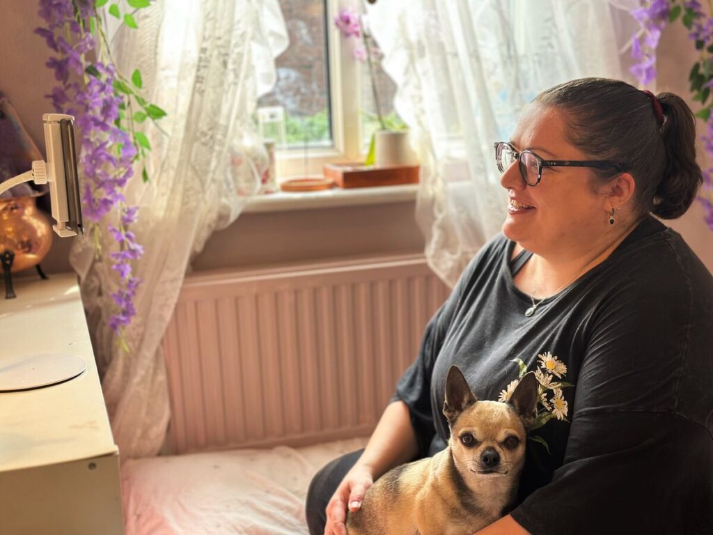 A woman sits at home with her small dog on her lap, taking part in an online session.