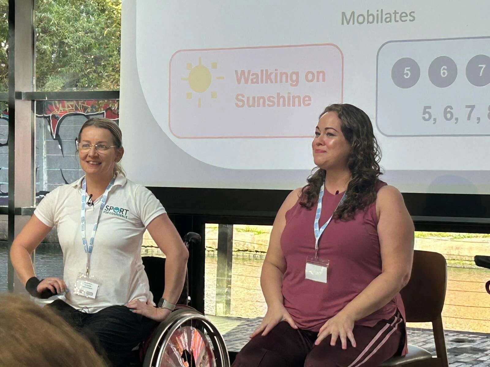 Leanne and Suzanne seated in front of a screen reading Walking on Sunshine, leading a movement session at the conference